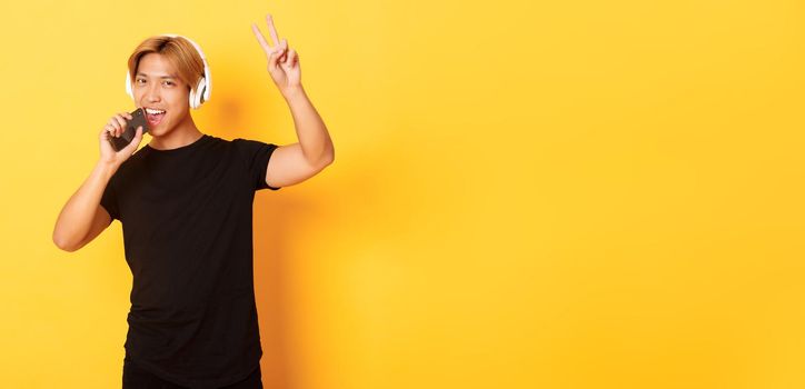 Sassy handsome asian guy in headphones, playing karaoke app, singing into mobile phone microphone, showing peace gesture, standing yellow background.