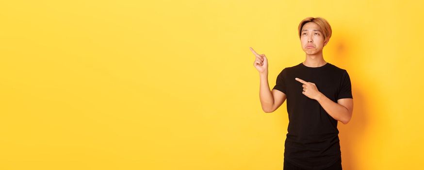 Portrait of pouting upset and funny asian guy pointing and looking upper left corner with regret, standing yellow background.