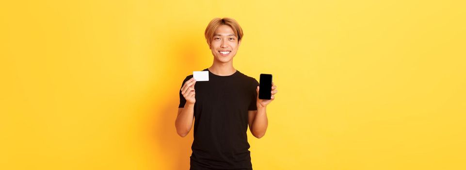 Portrait of handsome young asian guy showing smartphone screen, banking app and credit card, standing yellow background and smiling.