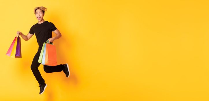 Full-length of happy attractive asian man jumping from happiness and carry shopping bags, standing yellow background.