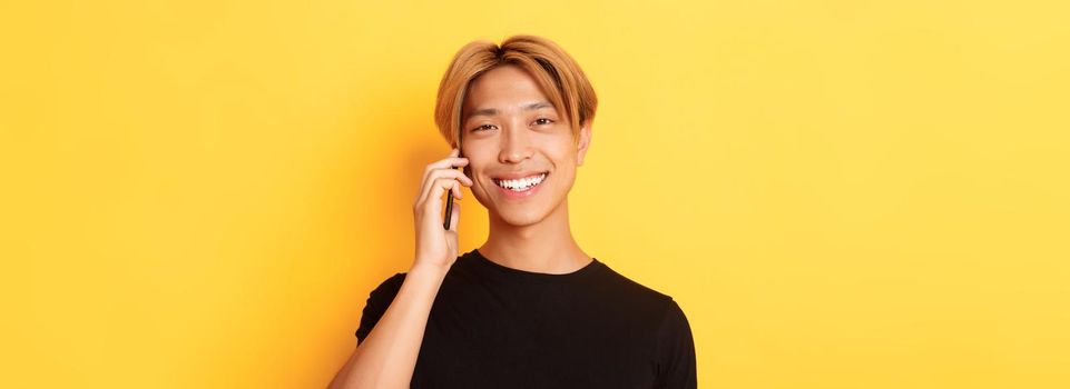 Close-up of handsome stylish korean guy smiling and talking on mobile phone, standing over yellow background.