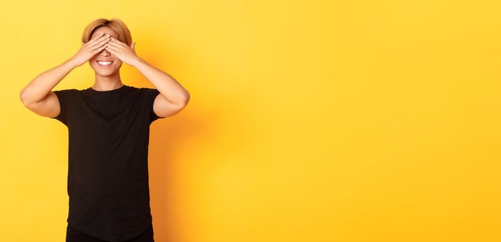 Portrait of excited happy asian guy, smiling and close eyes with hands, standing yellow background.