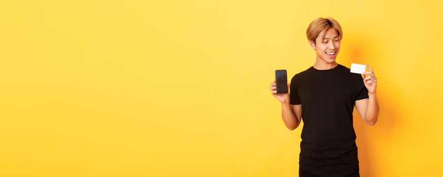 Handsome satisfied asian guy holding credit card and showing smartphone screen, smiling pleased, standing yellow background.