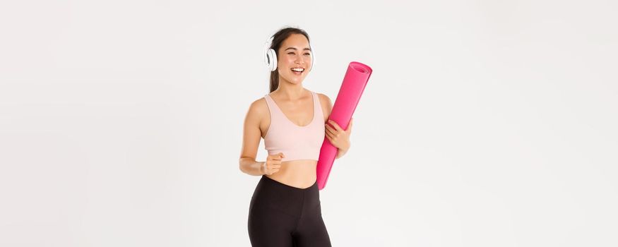 Sport, wellbeing and active lifestyle concept. Cheerful smiling asian fitness girl in headphones and sportswear going to gym with rubbermat, hurry up for yoga classes excited, white background.