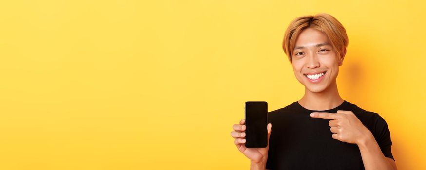 Cheerful attractive korean guy pointing finger at mobile phone screen, smiling pleased, standing over yellow background.