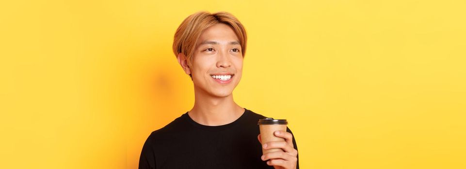 Close-up of relaxed and happy stylish asian blond guy, smiling and looking left dreamy while drinking coffee from takeaway cup, standing over yellow background.