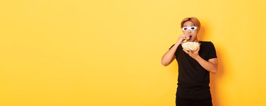 Impressed and thrilled asian guy attend cinema, watching movie or tv series in 3d glasses, eating popcorn and looking shocked, standing yellow background.