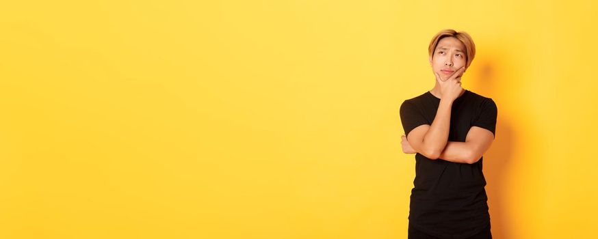 Portrait of thoughtful attractive guy in black t-shirt looking upper left corner. Young man pondering idea, standing yellow background.