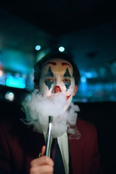 Guy in suit and tie with the Joker makeup puffs smoke out of his mouth. Autumn holiday. All Saints' Night. Halloween party in nightclub with decorations and scary costumes.