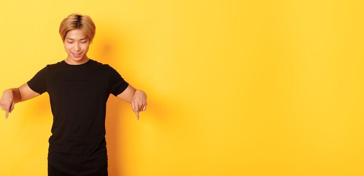 Portrait of curious happy blond asian guy, looking and pointing fingers down with pleased smile, yellow background.