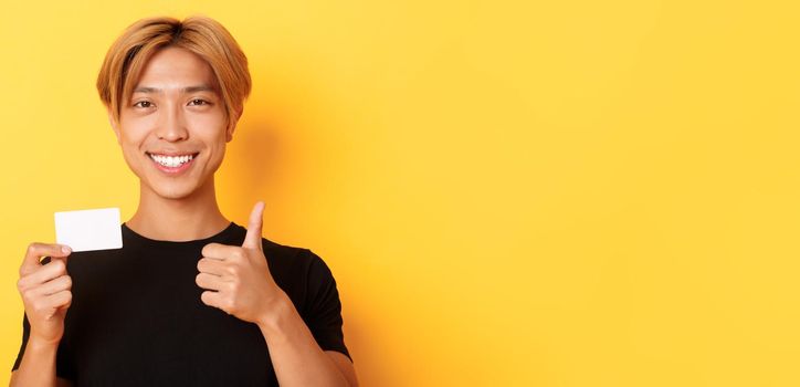 Close-up of satisfied asian guy recommend bank, showing credit card and standing over yellow background.