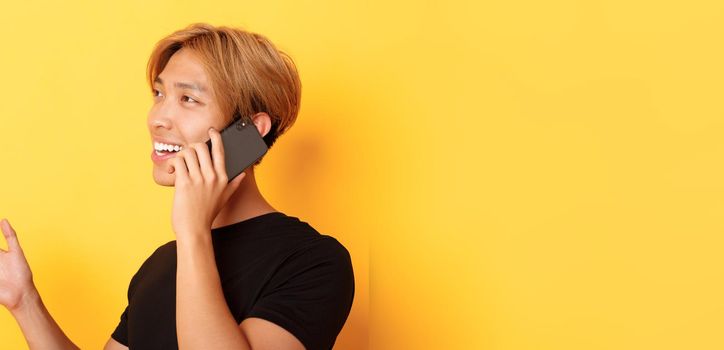 Close-up of attractive korean guy smiling and talking on mobile phone happy, turning left, standing yellow background.