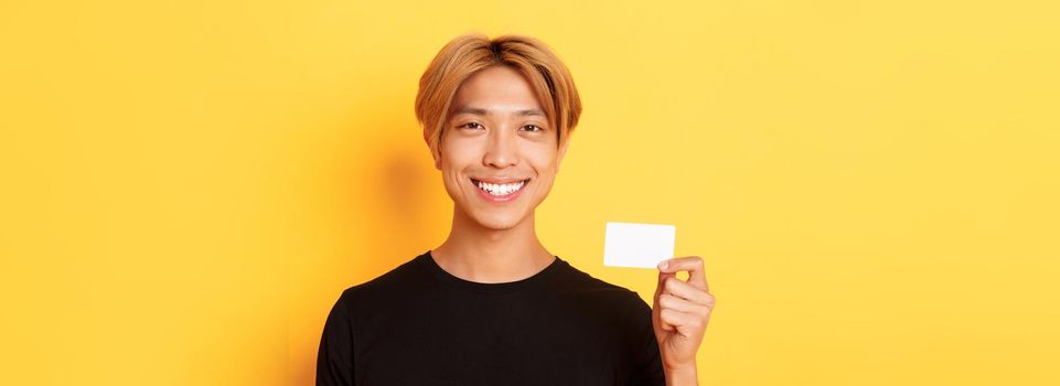 Close-up of handsome pleased asian guy with fair hair, smiling happy and showing credit card over yellow background.