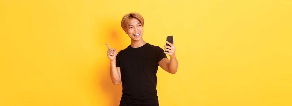 Portrait of handsome smiling asian man having video call on smartphone and pointing finger at something, standing yellow background.