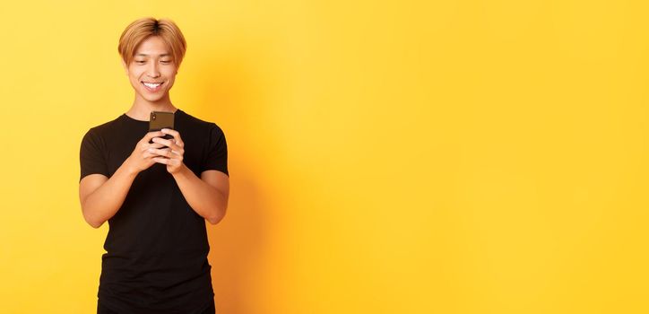 Portrait of handsome stylish asian guy with blond hair, using mobile phone and smiling, yellow background.