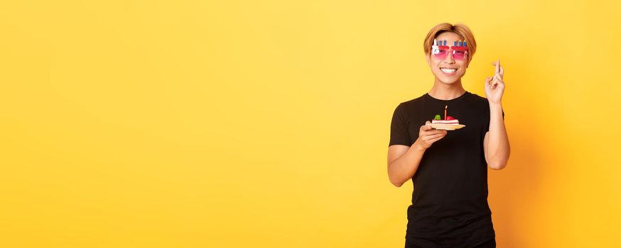 Portrait of hopeful attractive asian birthday guy, celebrating in funny party glasses, holding cake and fingers crossed while making b-day wish, yellow background.