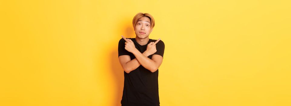 Indecisive handsome guy shrugging puzzled and pointing fingers sideways, standing yellow background.