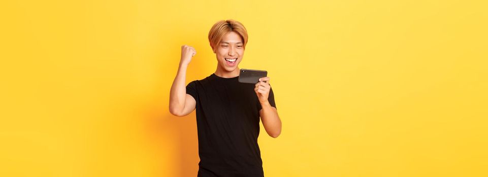 Portrait of stylish handsome asian guy playing smartphone game, looking amused at mobile screen and rejoicing from winning, standing yellow background.