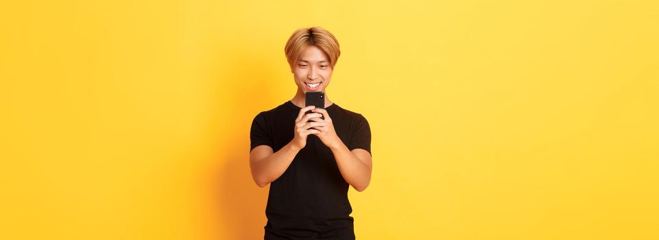 Portrait of handsome stylish asian guy with blond hair, using mobile phone and smiling, messaging in smartphone app, yellow background.