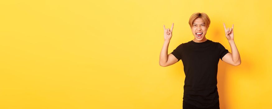 Portrait of happy handsome asian man having fun and partying, showing rock-n-roll gesture, standing yellow background.