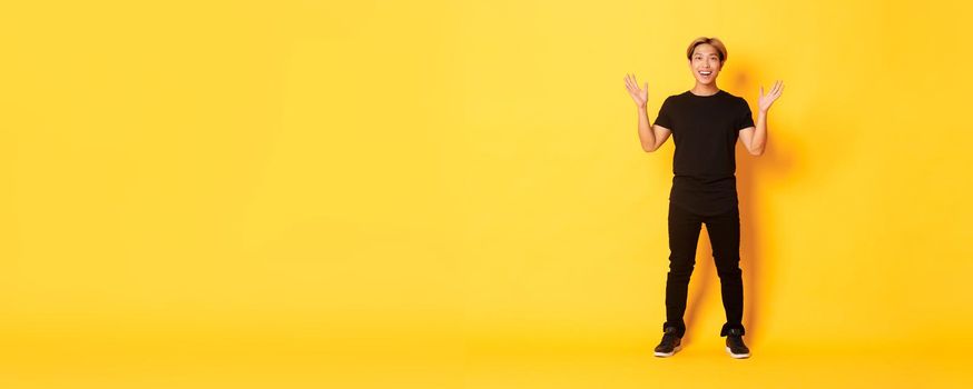 Full-length of surpirsed and excited, smiling blond asian guy raising hands up amazed, yellow background.