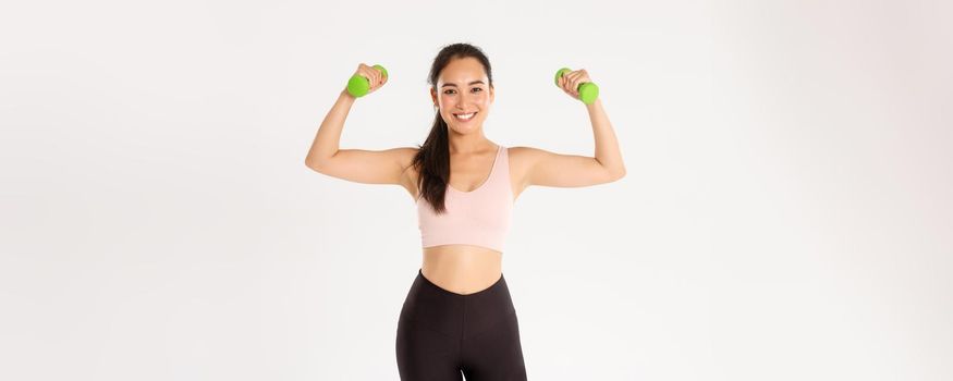 Fitness, healthy lifestyle and wellbeing concept. Portrait of strong and happy female athlete, asian girl workout at home during coronavirus, lifting dumbbells to gain muscles, white background.