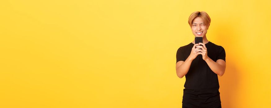 Stylish handsome asian guy taking picture on smartphone and smiling, photographing with mobile phone, standing yellow background.