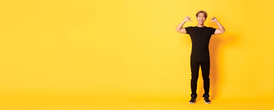 Confident attractive blond asian guy, smiling sassy and pointing at himself, standing over yellow background.