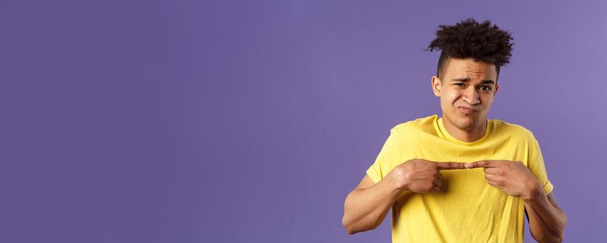 Close-up portrait of shy and modest young silly hispanic man trying say something but being too insecure, grimacing and frowning, look timid, two fingers touching pose, purple background.