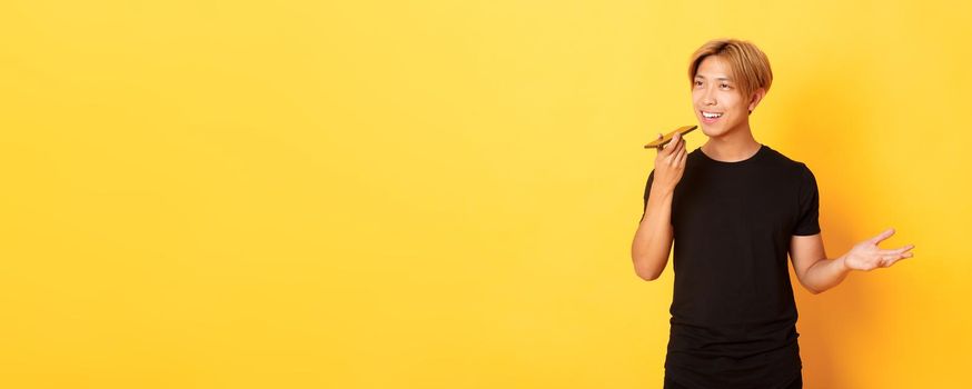 Portrait of stylish handsome korean guy with blond hair record voice message on mobile phone, holding smartphone near mouth and speaking, standing yellow background.