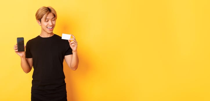 Handsome satisfied asian guy holding credit card and showing smartphone screen, smiling pleased, standing yellow background.