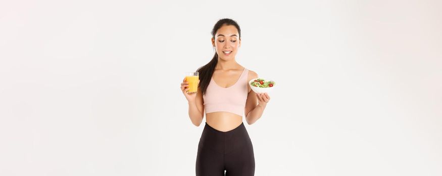 Sport, wellbeing and active lifestyle concept. Smiling healthy and slim brunette asian girl like fitness, going to gym and being on diet, holding salad with orange juice, standing white background.