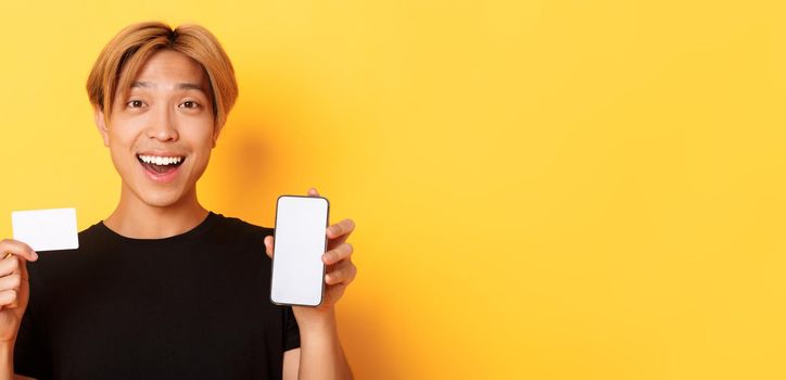 Amazed happy asian guy showing credit card and smartphone screen, smiling fascinated, standing yellow background.