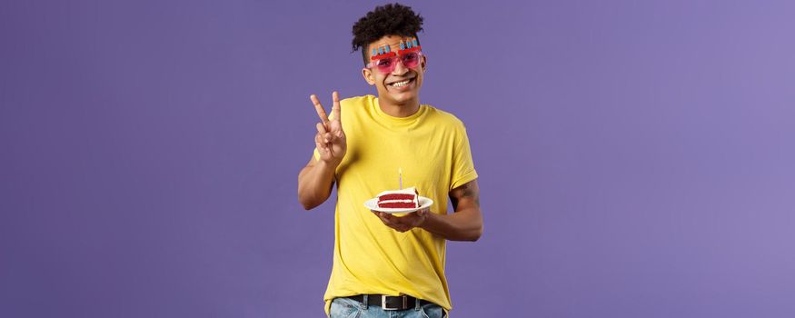 Celebration, party and holidays concept. Portrait of lovely charismatic hispaic guy in b-day glasses, celebrating his day, show peace sign photographing with birthday cake and lit candle, smiling.