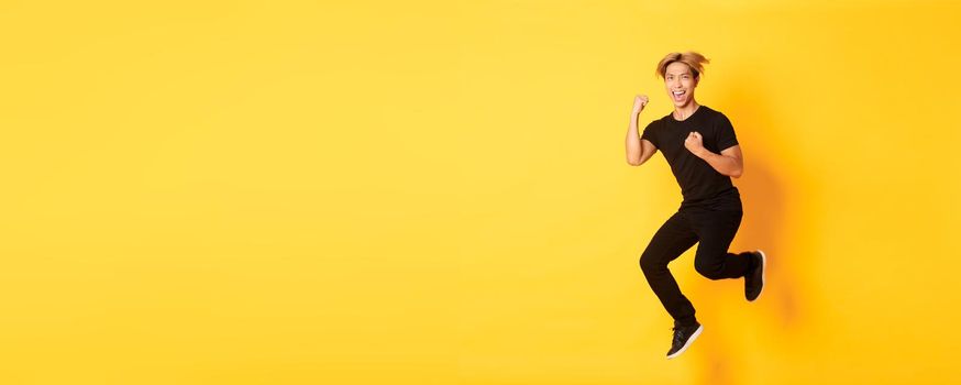 Full-length of happy attractive asian guy in black clothes jumping and celebrating victory, achieve goal, standing yellow background, triumphing.