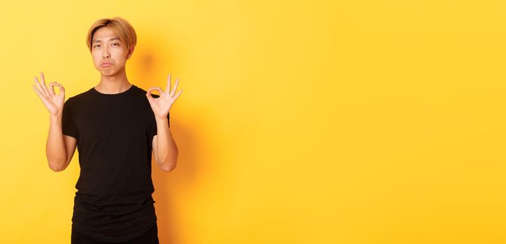 Impressed asian stylish guy with blond hair, showing okay gesture and praise something good, standing yellow background.