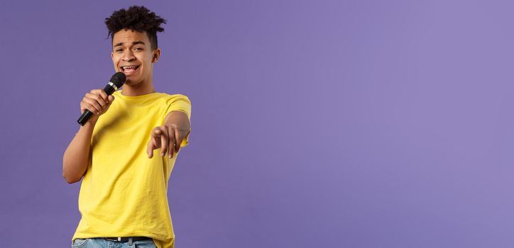 This song is for you. Portrait of romantic carefree hispanic man singing karaoke, pointing at camera as dedicate his performance, holding microphone, standing purple background.