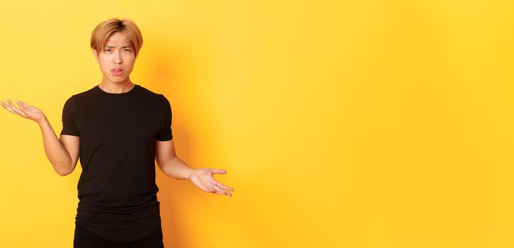 Portrait of confused and annoyed asian guy spread hands sideways, cant understand something, standing yellow background.