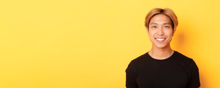 Close-up of handsome blond asian guy in black t-shirt, smiling happy at camera, standing over yellow background.