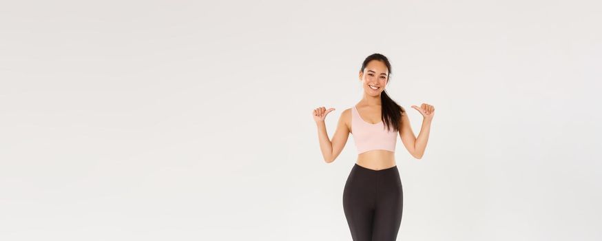 Full length of professional fitness coach, asian girl athelte pointing at herself and smiling happy, workout in gym, gaining perfect body with training, wearing sportswear, white background.