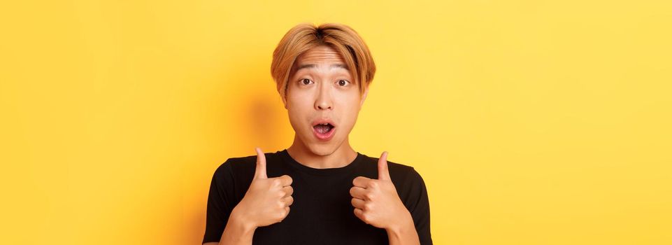 Close-up of impressed and excited handsome asian guy showing thumbs-up and looking amazed, standing over yellow background.
