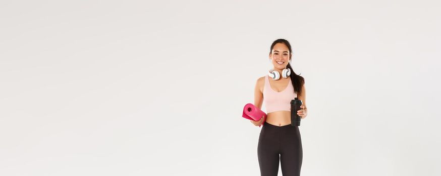 Full length of beautiful asian girl going for yoga classes, female athlete with headphones and water bottle smiling as carry fitness exercises rubber mat for her training in gym, workout concept.