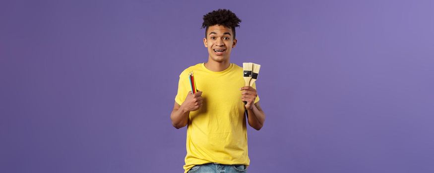 Portrait of upbeat young creative man, artist starting art courses, want to learn how to draw, holding colored pencils and brushes, standing enthusiastic over purple background.