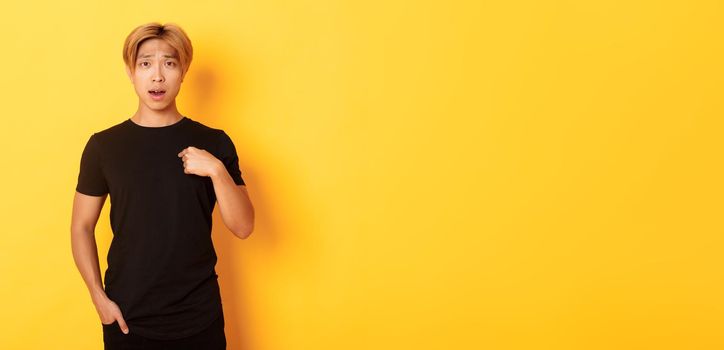 Portrait of confused and sad asian blond guy, frowning upset and pointing at himself, standing yellow background.