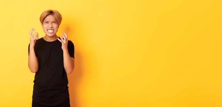 Portrait of angry and pissed-off asian blond guy, clenching hands furious and grimacing, standing over yellow background.