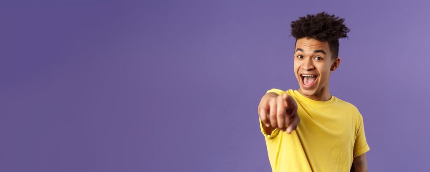 Close-up portrait of upbeat, amazed hispanic man with dreads, young student pointing finger at camera and laughing, recognize someone familiar, standing purple background.