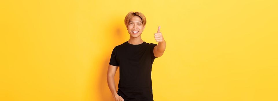 Portrait of satisfied asian man with blond hair, standing over yellow background and showing thumbs-up.