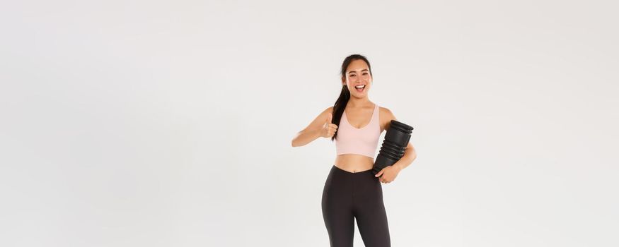 Full length of satisfied smiling asian female training coach, fitness girl in sportswear looking pleased, showing thumbs-up, using foam roller after workout to reduce soreness and ache in muscles.