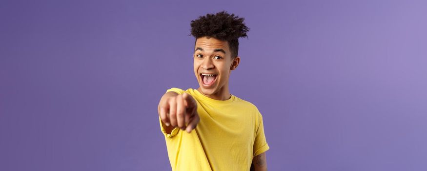 Close-up portrait of upbeat, amazed hispanic man with dreads, young student pointing finger at camera and laughing, recognize someone familiar, standing purple background.