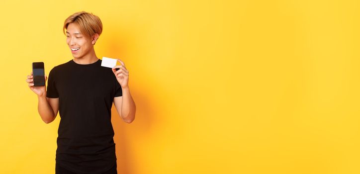 Portrait of pleased handsome asian guy, showing smartphone screen and credit card, looking at mobile phone display, yellow background.